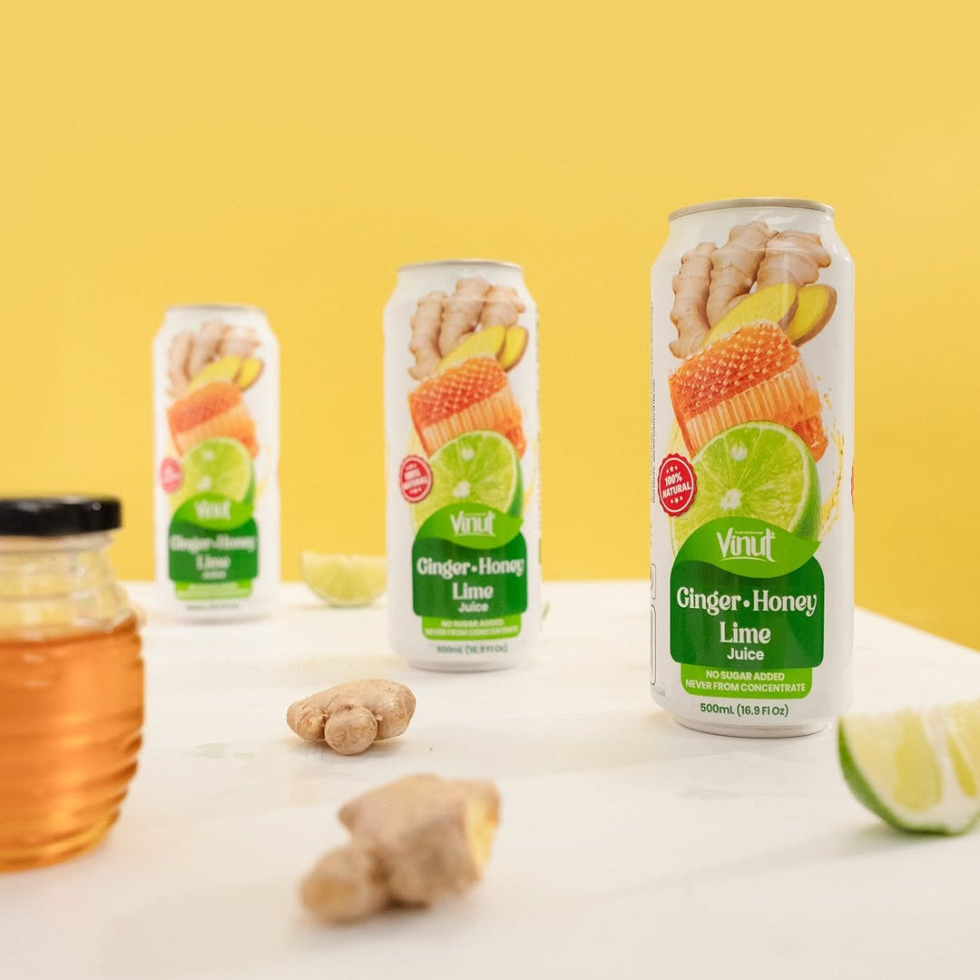 Three VINUT Ginger Honey Lime juice cans with fresh ginger, honey and lime on a yellow background